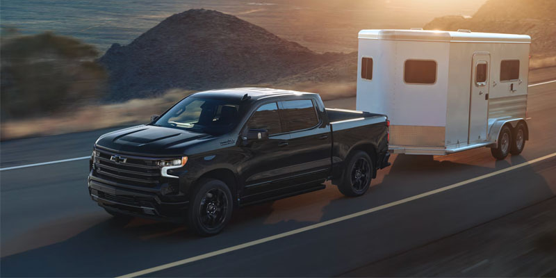 Black Chevrolet pickup truck towing a white trailer along a coastal road at sunset
